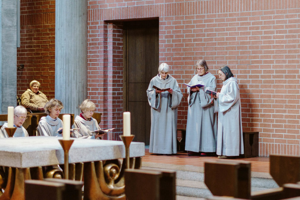 Drei Schwestern in weißer Ordenstracht singen im stehen, während die anderen im Chorgestühl sitzen.